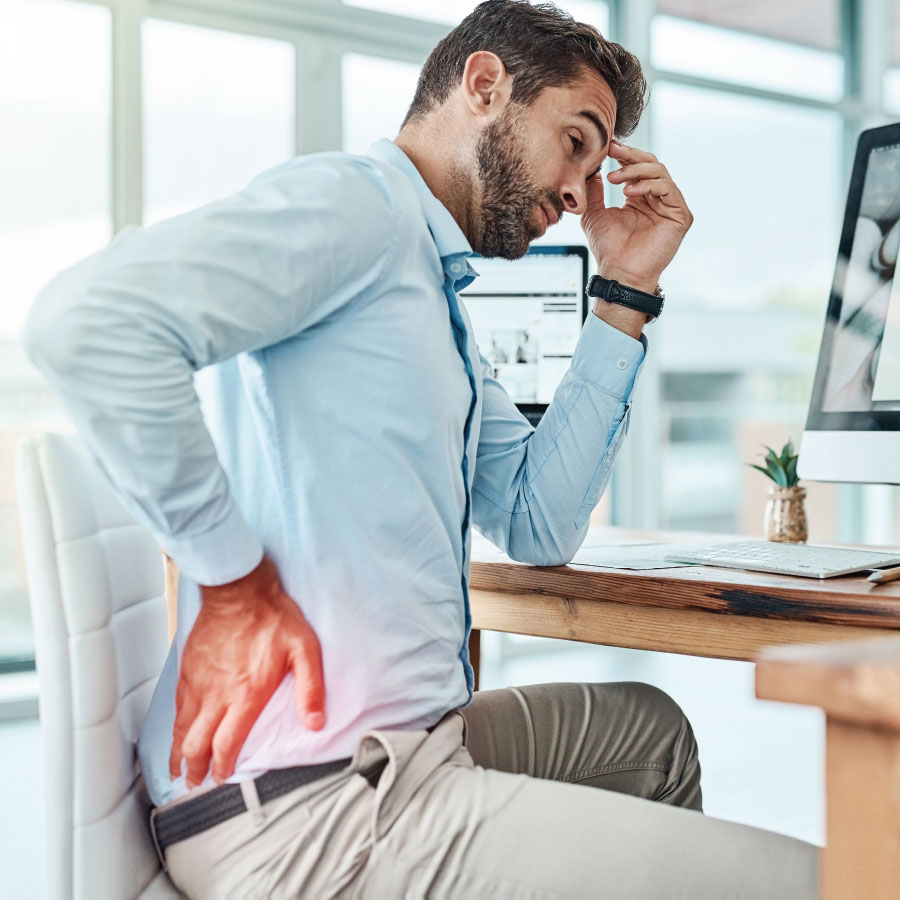 A man in an office setting sits at a desk, looking pained. He holds his lower back with one hand, indicating discomfort or back pain.
