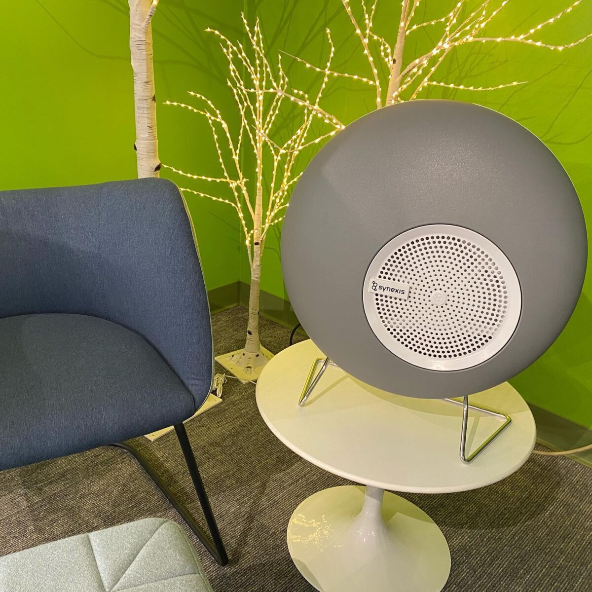 A modern room with a gray speaker on a small round table, a stylish blue chair, and lit decorative white branches. The green wall adds vibrancy.