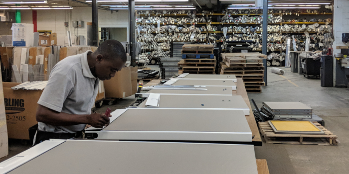 A person in a warehouse, focused on assembling white boards. Shelves filled with fabric rolls are in the background, conveying a busy, productive atmosphere.