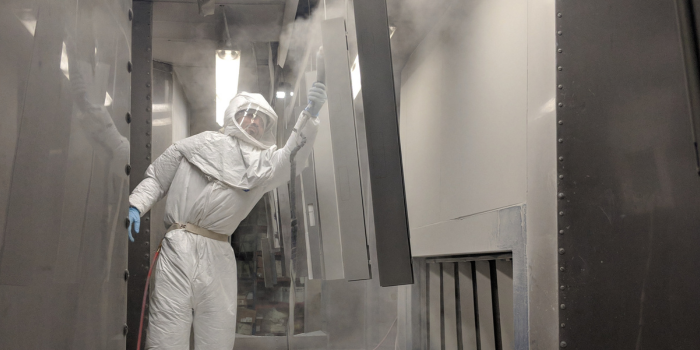 A person in protective gear spray paints a large object inside a workshop. The room is filled with mist, and the scene has a focused, industrious tone.