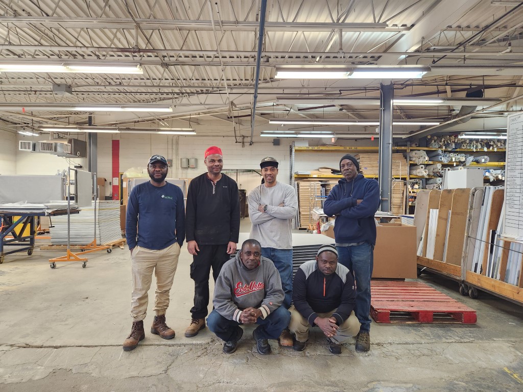 Group of six people in a warehouse, wearing casual work attire. Two kneel in front, four stand behind. The atmosphere is casual and friendly.