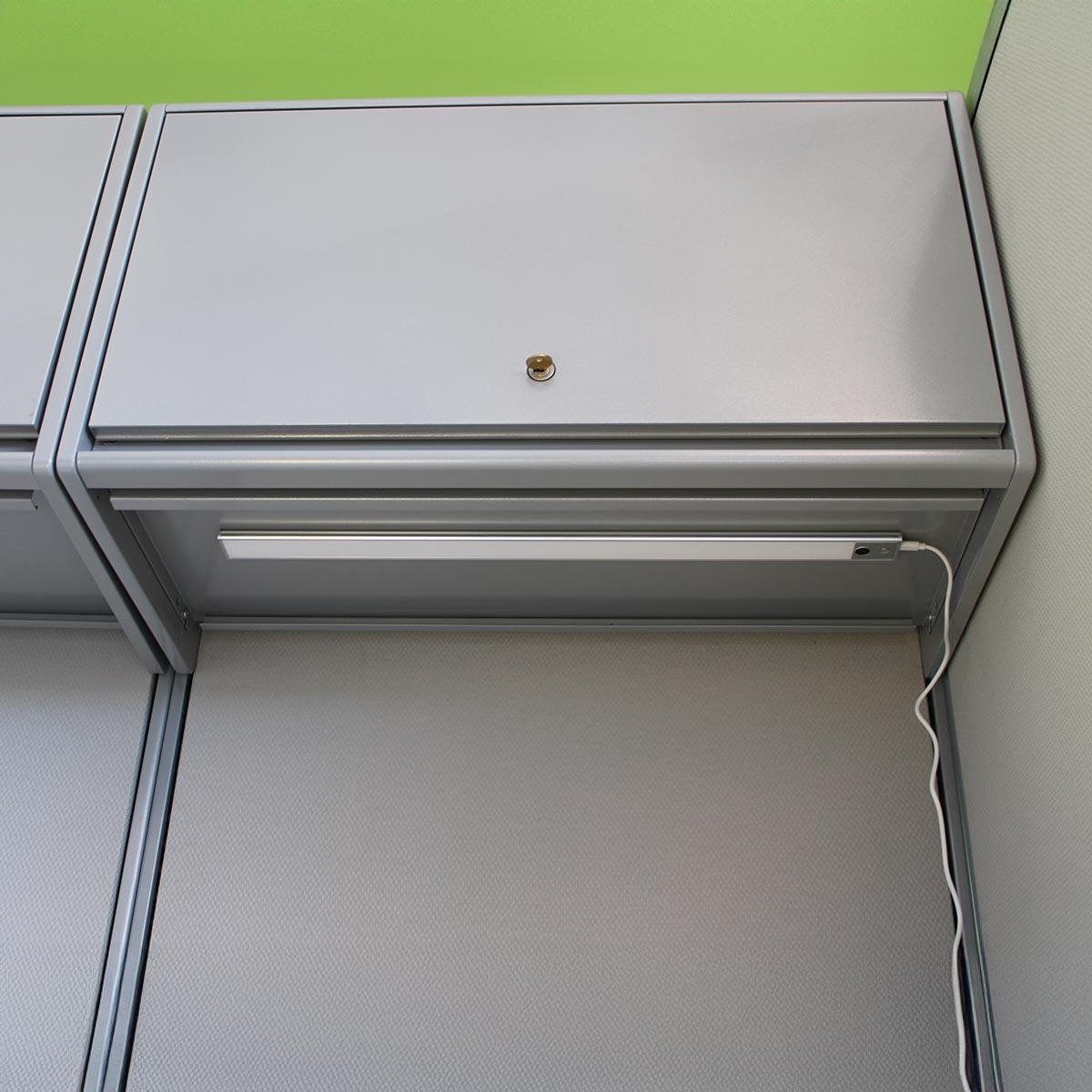 Office cubicle with closed top cabinet, under-cabinet light, and visible power cord. Lime green wall strip above adds contrast to the neutral tones.