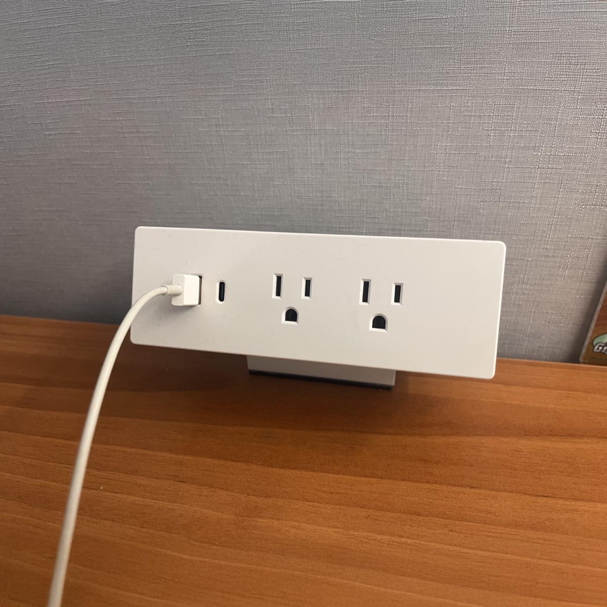 A white power strip with three outlets, one occupied by a plugged-in white cable, sits on a wooden desk against a gray wall, conveying utility.