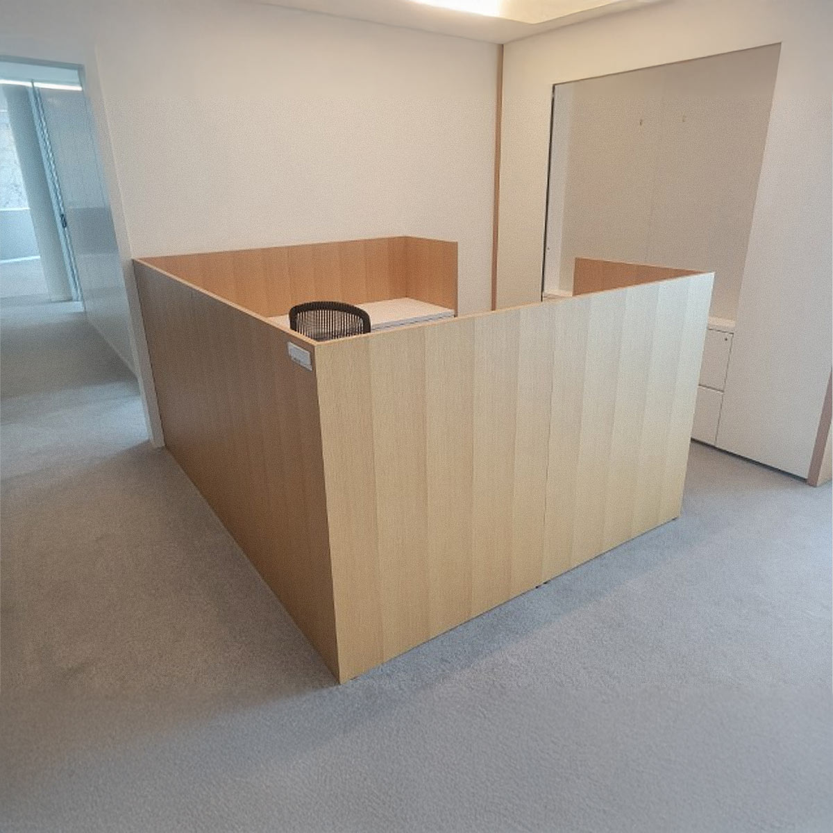 Wooden cubicle set in a bright office space with gray carpet. Inside is an empty desk and a black mesh chair, creating a minimalist, professional atmosphere.