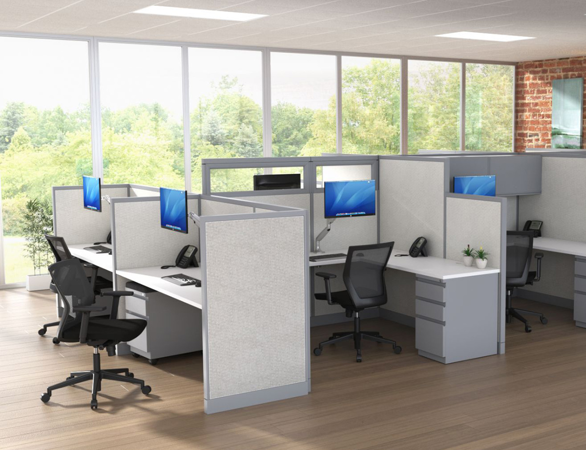Modern office with gray cubicles, black chairs, and computers on desks. Large windows show a green outdoor view, creating a bright, airy feel.