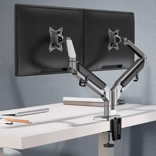 A dual monitor stand with adjustable arms holds two screens on a white desk in an office setting. Notebooks and pencils are neatly arranged nearby.