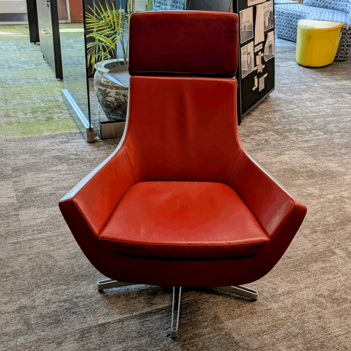 A modern red swivel chair with a high backrest sits in an office setting. The room features gray carpeting, a potted plant, and a yellow stool.