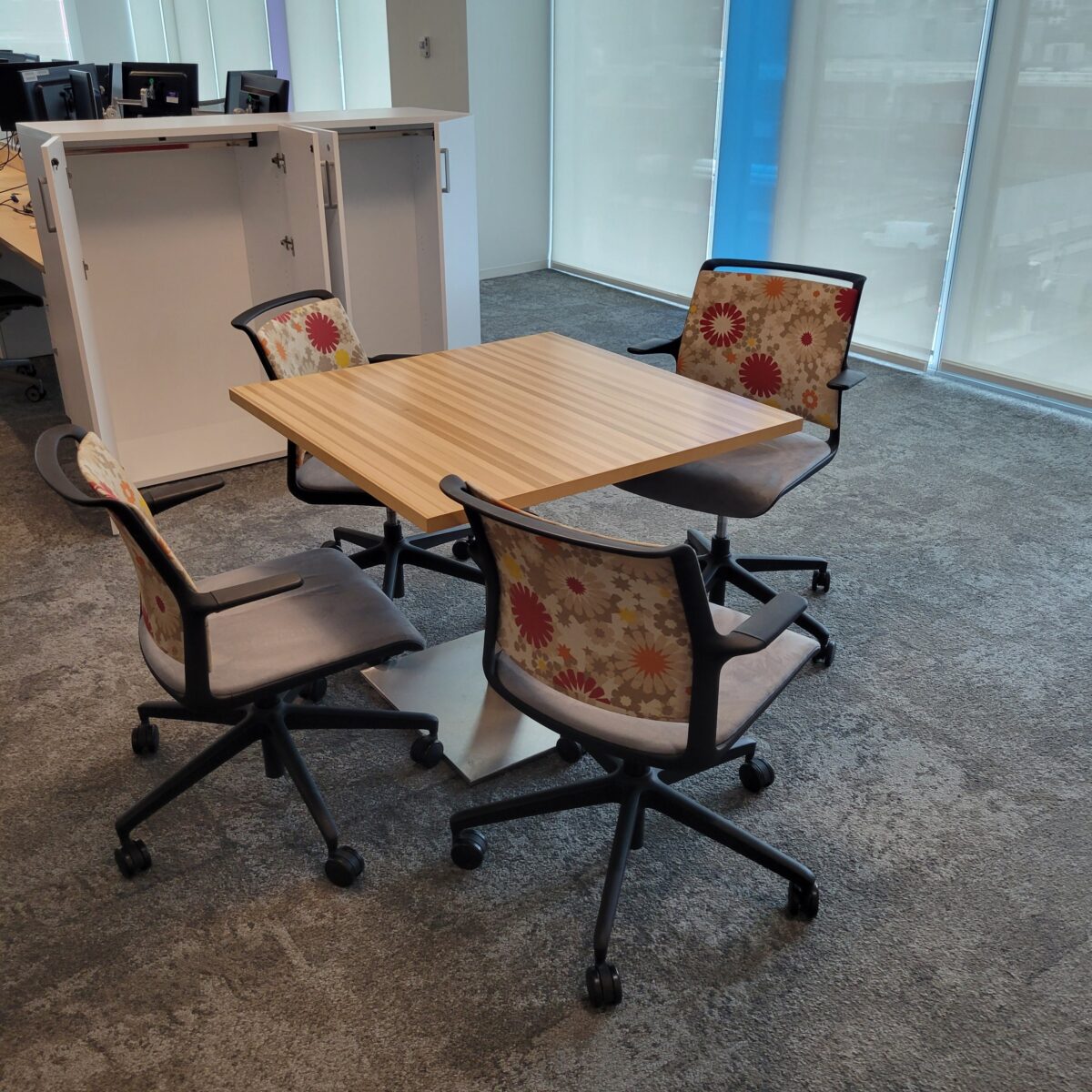 A modern office meeting area features a wooden square table surrounded by four wheeled chairs with colorful floral-patterned backs, set on a gray carpet.