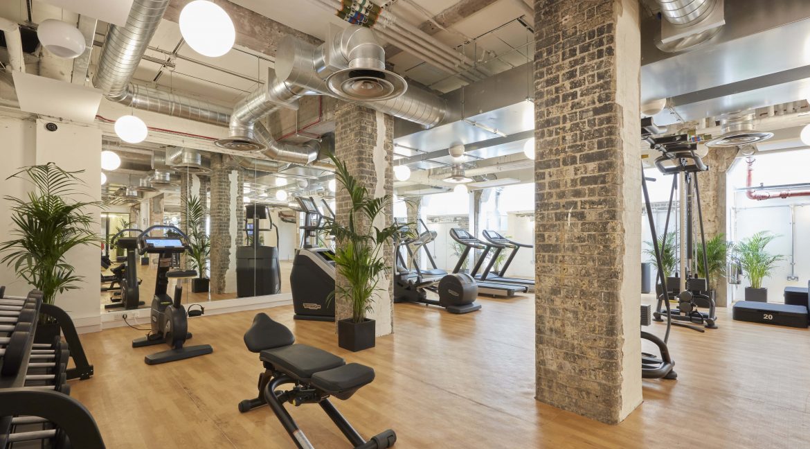 Spacious gym with wooden floors, exposed brick pillars, and large mirrors. Equipped with treadmills, weight benches, stationary bikes, and plants, creating an airy, modern vibe.