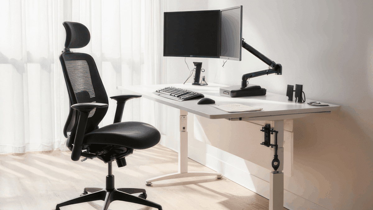 Modern office setup with an ergonomic black chair and a white desk featuring a dual monitor arm, keyboard, and mouse. Sunlight filters through sheer curtains.