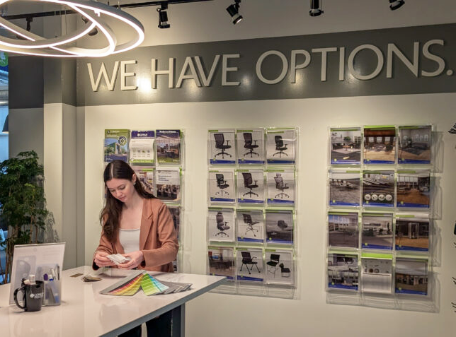 A woman examines color swatches on a table in a showroom with "WE HAVE OPTIONS" on the wall. Chair brochures are displayed behind her. Modern, bright setting.