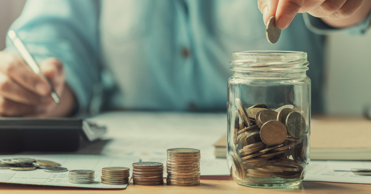 A person in a blue shirt places a coin into a jar full of coins, surrounded by stacks of coins and financial documents. The tone is focused and contemplative.