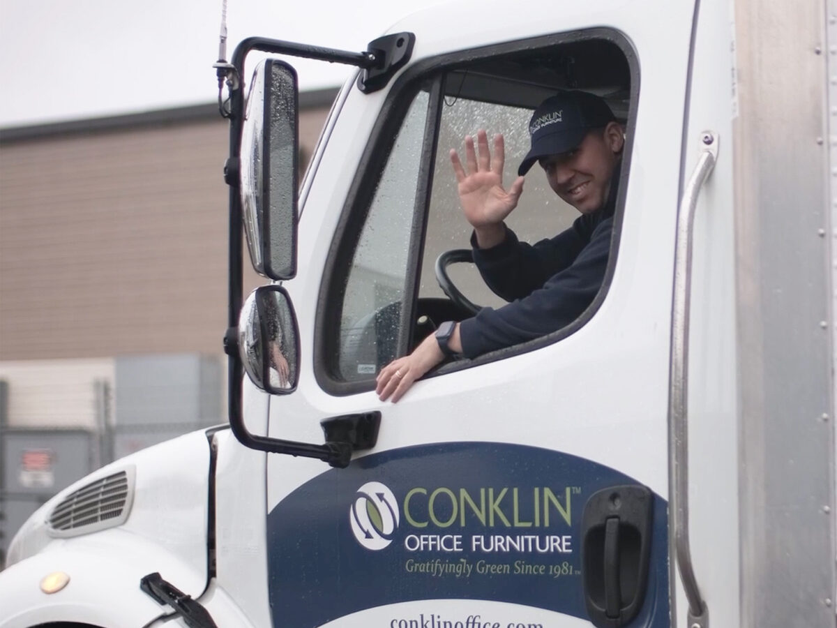Man waves smiling from inside a Conklin Office Furniture truck cab. The truck is parked outside a building, conveying a friendly and professional tone.